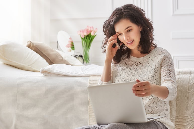A woman working on her computer in front of a bed.